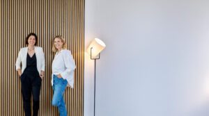Die Gründerinnen von Make It Matter, Lisa und Christina, stehen in einem modernen Büro vor einer Holzstrukturwand – Kommunikationsberatung für Unternehmen mit Fokus auf Strategie, Wirkung und Wachstum.