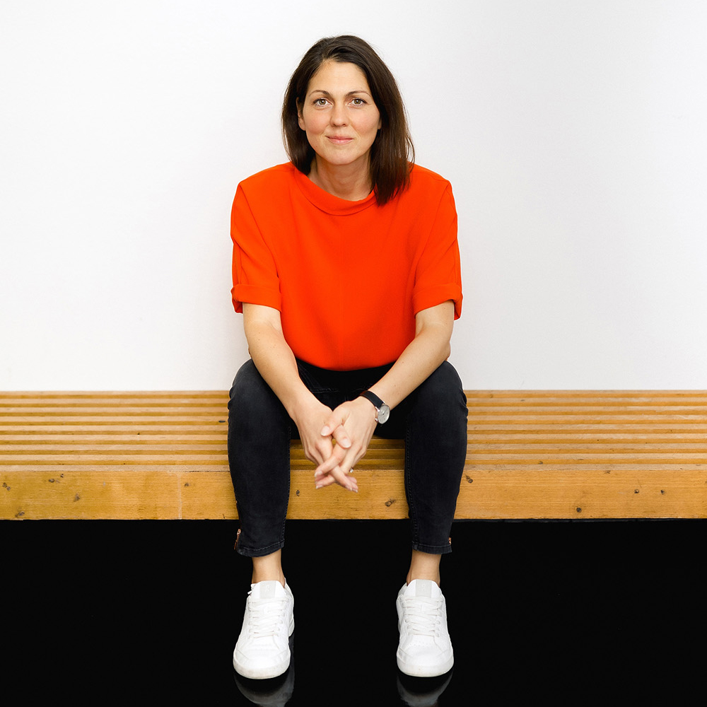 Lisa, Co-Founderin von Make It Matter, Kommunikationsberaterin und Strategin für Markenpositionierung und LinkedIn-Kommunikation, sitzt auf einer Holzbank und blickt freundlich in die Kamera