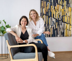Die beiden Gründerinnen von Make It Matter, Lisa und Christina, sitzen gemeinsam in einem modernen Büro und lächeln – Symbol für Kommunikationsberatung, Teamarbeit und strategische Zusammenarbeit auf Augenhöhe.