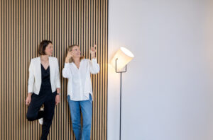 Kontakt - Zwei Kommunikationsberaterinnen von Make It Matter stehen vor einer modernen Holzstrukturwand und lächeln – Symbol für persönliche, offene Beratung und Kommunikation auf Augenhöhe.